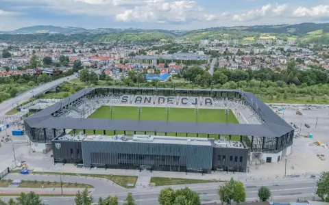 Nowoczesny stadion Sandecji z zieloną murawą i trybunami, otoczony miastem i wzgórzami.