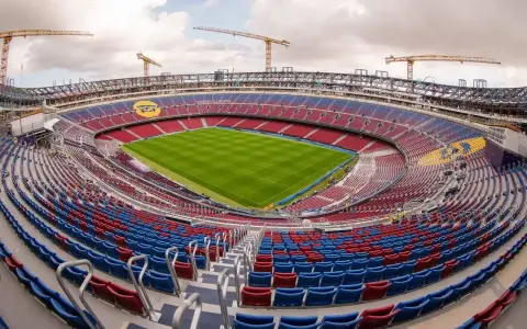 Widok na budowę stadionu, gdzie powstaje nowe Camp Nou. Żurawie górują nad trybunami w kolorach czerwonym i niebieskim, a murawa lśni zielenią.