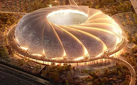 Futurystyczny stadion, który może być najładniejszy na świecie, z falującym dachem i oświetlonymi detalami, otoczony zielenią i tłumem ludzi.