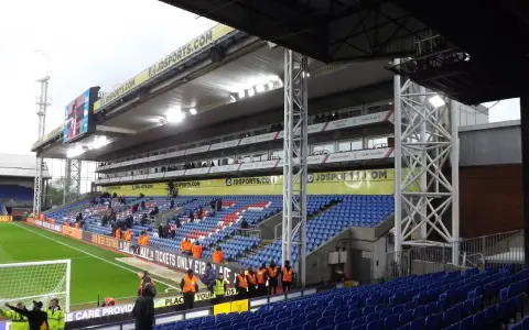 Puste trybuny stadionu Selhurst Park, z niebieskimi i czerwonymi siedzeniami, zieloną murawą i wielkim telebimem.
