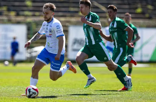 Piłkarz w białej koszulce z niebieskimi spodenkami prowadzi piłkę na boisku. W tle zawodnicy w zielonych strojach. Mecz forbet 4 liga warmińsko-mazurska.