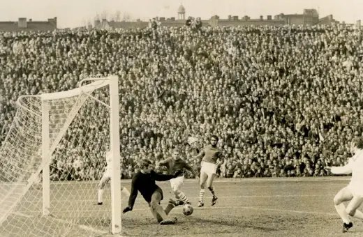 Byli piłkarze Lecha Poznań w akcji na pełnym stadionie. Bramkarz broni, napastnik strzela, a tłum kibiców obserwuje.