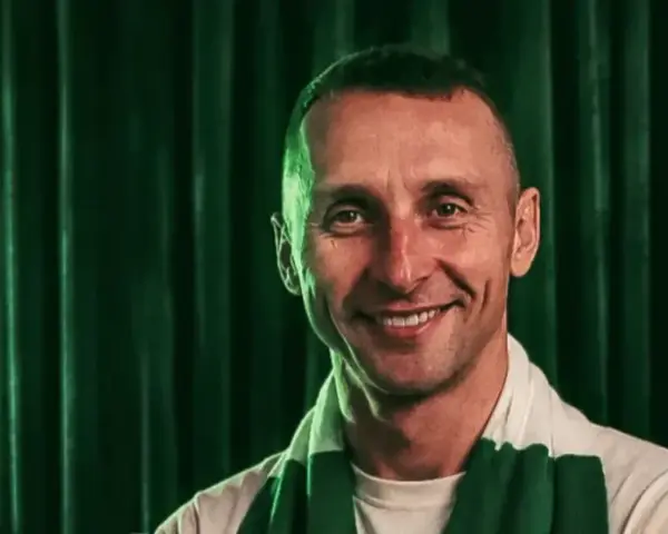 Uśmiechnięty trener Lechii Gdańsk w szaliku klubowym na tle zielonej kurtyny.