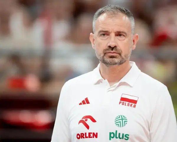 Skupiony trener siatkarzy reprezentacji Polski w białej koszulce z godłem narodowym i logotypami sponsorów.