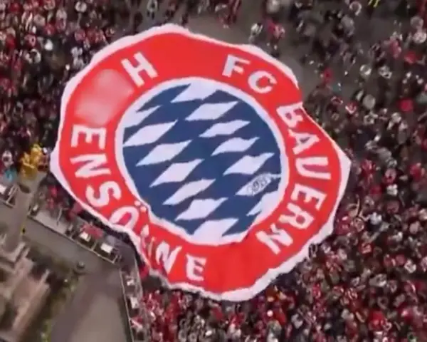 Ogromna flaga z herbem FC Bayern Monachium niesiona przez tłum kibiców.