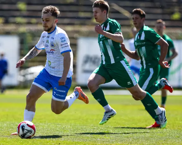 Piłkarz w białej koszulce z niebieskimi spodenkami prowadzi piłkę na boisku. W tle zawodnicy w zielonych strojach. Mecz forbet 4 liga warmińsko-mazurska.