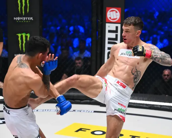 Zacięta walka na KSW 115 karta walk. Zawodnik w białych spodenkach wykonuje wysokie kopnięcie, przeciwnik blokuje je rękawicą.