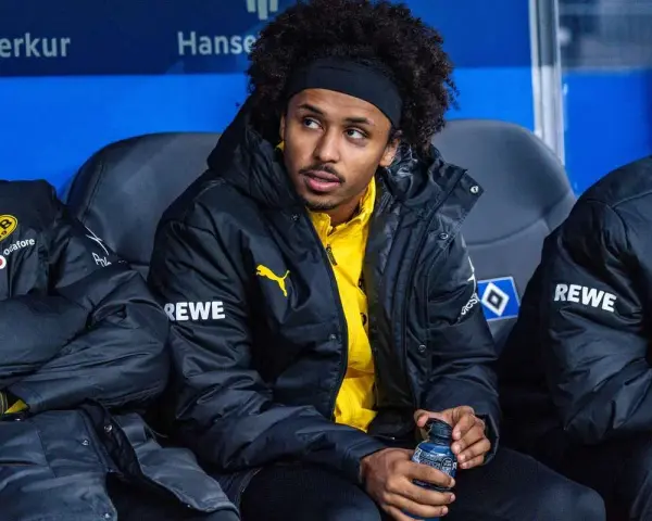 Piłkarz Adeyemi w czarnej kurtce z żółtym logo BVB i napisem REWE, siedzi na ławce rezerwowych, trzymając butelkę z napojem.