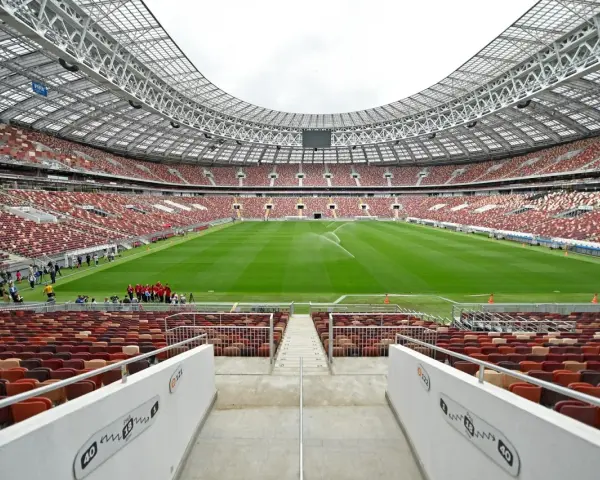 Pusta trybuna moskiewskiego stadionu z rzędem czerwonych i brązowych siedzeń. Na boisku pracują zraszacze.