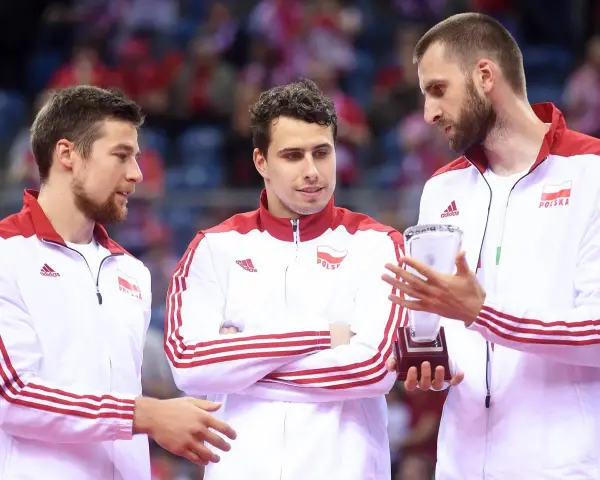 Trzech siatkarzy w strojach z biało-czerwoną flagą Polski. Jeden z nich trzyma puchar, symbolizujący, kto jest aktualnym mistrzem świata w siatkówce.