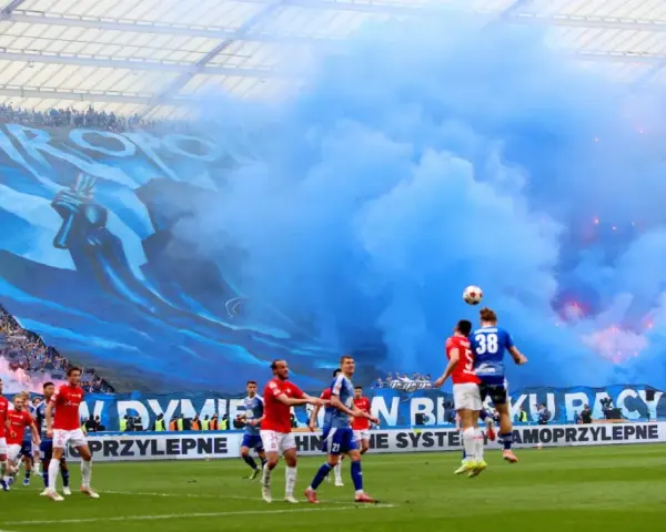 Mecz Ruch Chorzów – Wisła Kraków. Niebieskie dymy i ogromna oprawa kibiców na trybunach. Piłkarze walczą o piłkę na murawie.