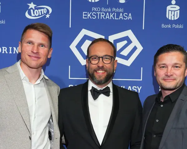 Trzech mężczyzn na tle PKO Bank Polski Ekstraklasa. Jeden z nich, w garniturze, uśmiecha się do obiektywu, być może jako jeden z przyszłych królowie strzelców ekstraklasy.