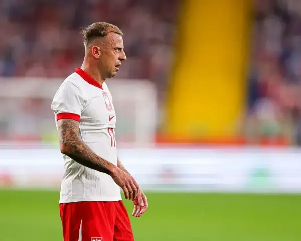 Kamil Grosicki w białej koszulce z orzełkiem i czerwonych spodenkach, gotowy do gry. Jego obecne drużyny to Polska i Pogoń Szczecin.