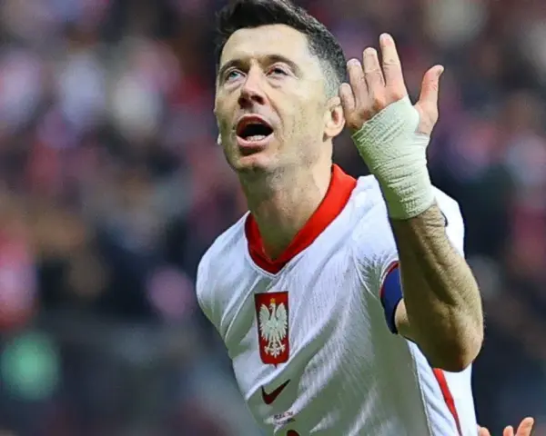 Robert Lewandowski w koszulce reprezentacji Polski, z bandażem na ręce, wznosi gest ku górze. Fani zastanawiają się, o której mecz piłki nożnej.