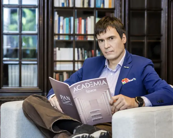 Mężczyzna w garniturze czyta magazyn "Academia Space" w bibliotece. W tle regały z książkami.