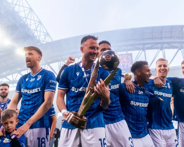 Piłkarze w niebieskich strojach świętują z pucharem i piłką na stadionie.