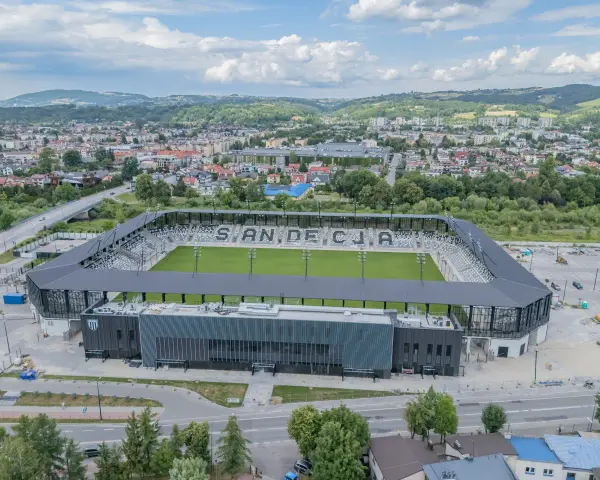 Nowoczesny stadion Sandecji z zieloną murawą i trybunami, otoczony miastem i wzgórzami.