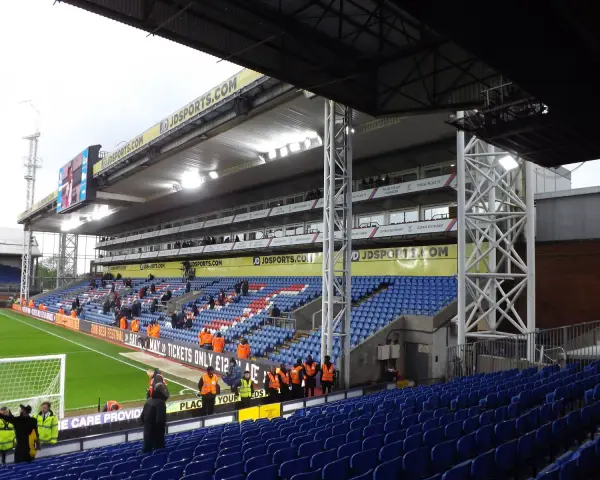 Puste trybuny stadionu Selhurst Park, z niebieskimi i czerwonymi siedzeniami, zieloną murawą i wielkim telebimem.