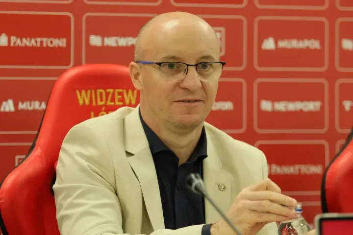 Nowy trener Widzewa w jasnej marynarce, siedzi na czerwonym fotelu z logo klubu.