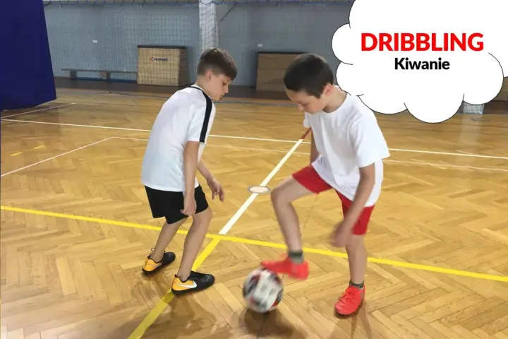 Dwóch chłopców ćwiczy drybling piłką nożną na sali gimnastycznej.