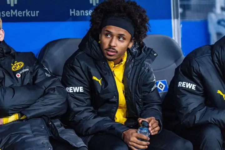 Piłkarz Adeyemi w czarnej kurtce z żółtym logo BVB i napisem REWE, siedzi na ławce rezerwowych, trzymając butelkę z napojem.