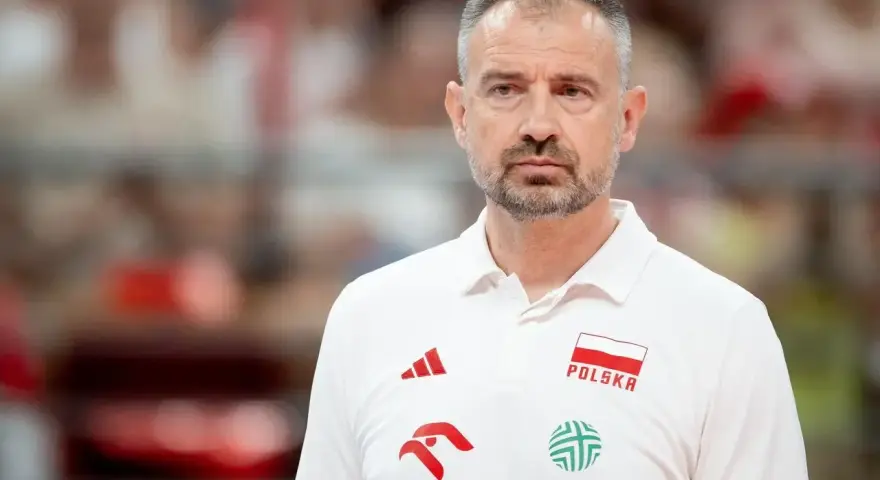 Skupiony trener siatkarzy reprezentacji Polski w białej koszulce z godłem narodowym i logotypami sponsorów.