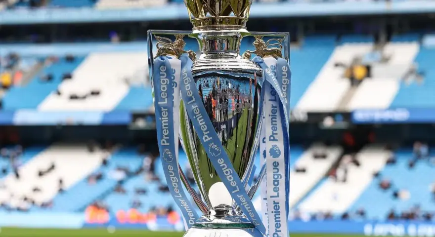 Trofeum za mistrzostwo Anglii, błyszczące i ozdobione wstążkami Premier League, stoi na tle stadionu.