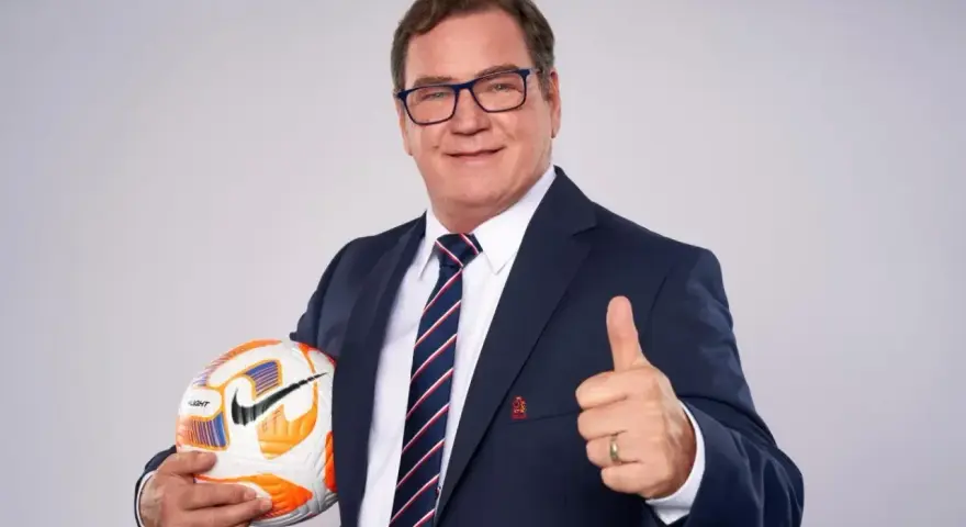 Uśmiechnięty trener reprezentacji Polski w garniturze trzyma piłkę i pokazuje kciuk w górę.