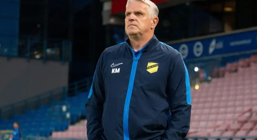 Trener z Krakowa, w granatowej kurtce z żółtym herbem, patrzy w dal na stadionie.