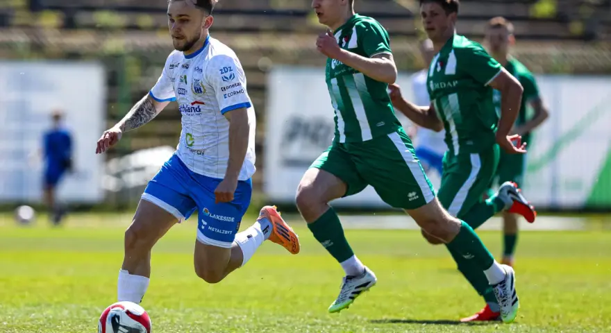 Piłkarz w białej koszulce z niebieskimi spodenkami prowadzi piłkę na boisku. W tle zawodnicy w zielonych strojach. Mecz forbet 4 liga warmińsko-mazurska.