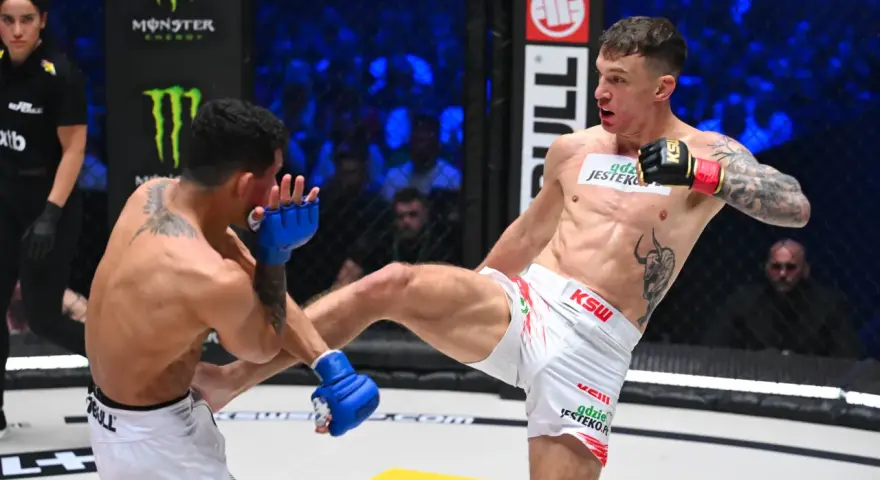 Zacięta walka na KSW 115 karta walk. Zawodnik w białych spodenkach wykonuje wysokie kopnięcie, przeciwnik blokuje je rękawicą.