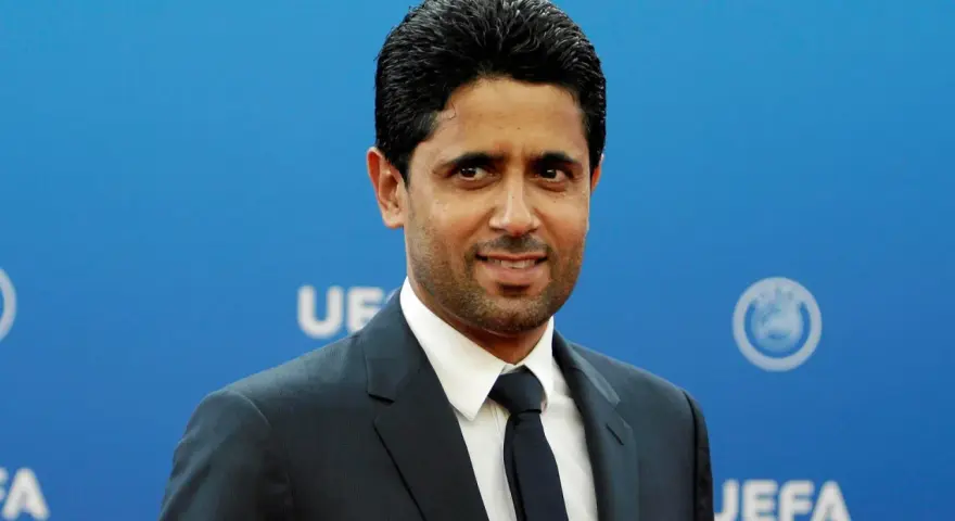 Nasser Al-Khelaifi w garniturze na tle niebieskiej ściany z logo UEFA.