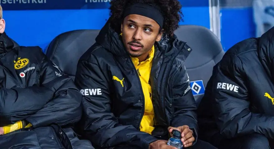 Piłkarz Adeyemi w czarnej kurtce z żółtym logo BVB i napisem REWE, siedzi na ławce rezerwowych, trzymając butelkę z napojem.