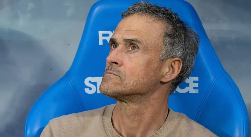 Luis Enrique, kto jest trenerem PSG, siedzi na niebieskiej ławce, zamyślony.