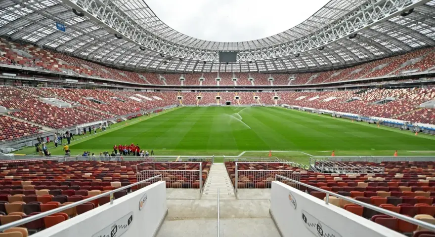 Pusta trybuna moskiewskiego stadionu z rzędem czerwonych i brązowych siedzeń. Na boisku pracują zraszacze.