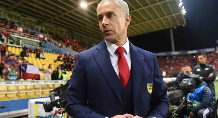 Sylvinho, trener w granatowym garniturze i czerwonym krawacie, stoi na tle trybun stadionu z flagą Chile.