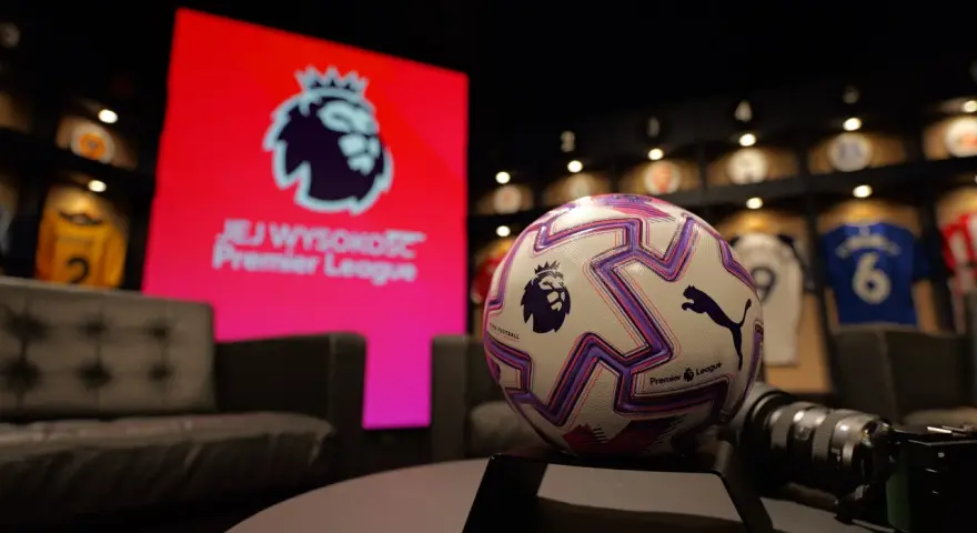 Piłka do gry w Premier League na tle ekranu z logo ligi. W tle widoczne koszulki piłkarskie.