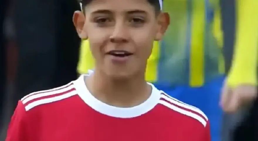 Młody piłkarz w czerwonej koszulce Manchester United, przypominający syna Cristiano Ronaldo. Jego wiek sugeruje, że to początki kariery.