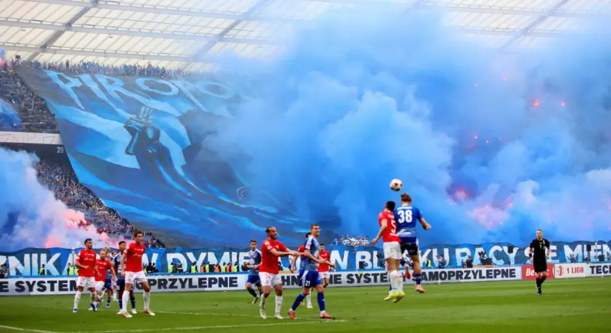 Mecz Ruch Chorzów – Wisła Kraków. Niebieskie dymy i ogromna oprawa kibiców na trybunach. Piłkarze walczą o piłkę na murawie.