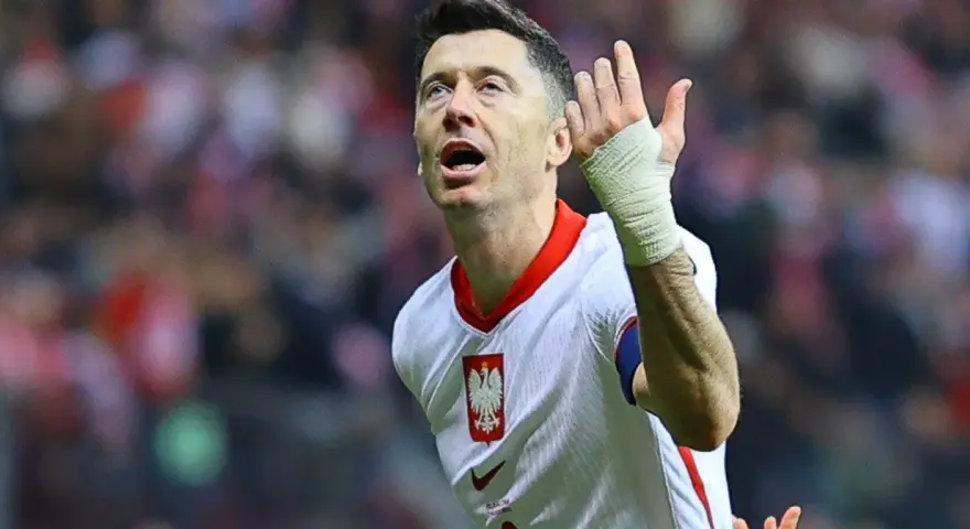 Robert Lewandowski w koszulce reprezentacji Polski, z bandażem na ręce, wznosi gest ku górze. Fani zastanawiają się, o której mecz piłki nożnej.
