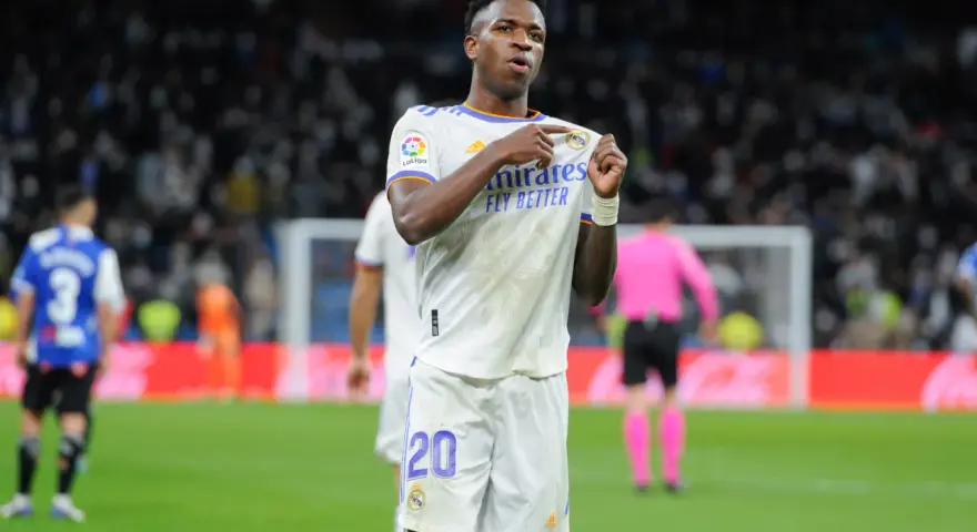 Vinicius Jr. w białej koszulce Realu Madryt z numerem 20, świętuje bramkę na tle trybun.
