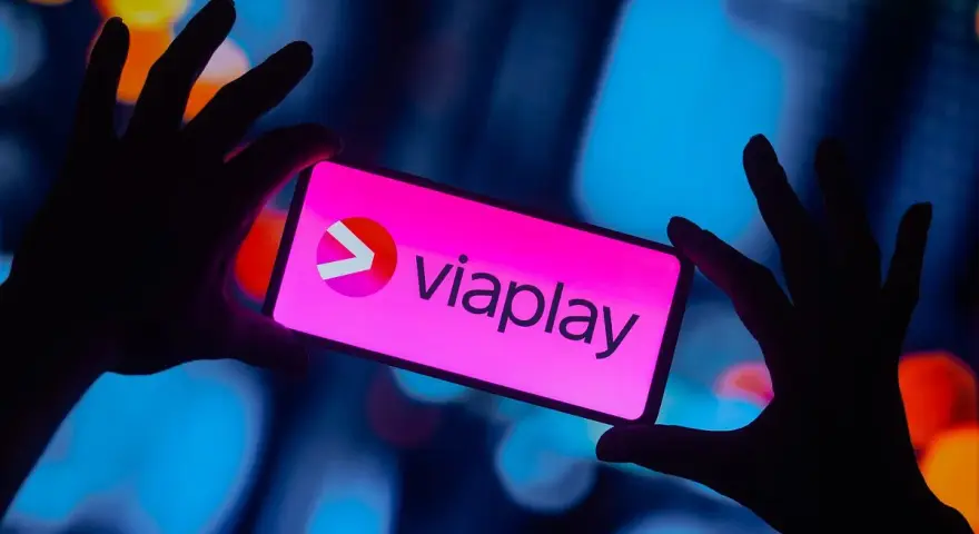 Dłonie trzymają smartfon z logo Viaplay. W tle rozmyte, kolorowe światła.