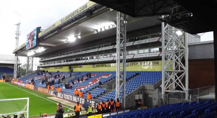Puste trybuny stadionu Selhurst Park, z niebieskimi i czerwonymi siedzeniami, zieloną murawą i wielkim telebimem.