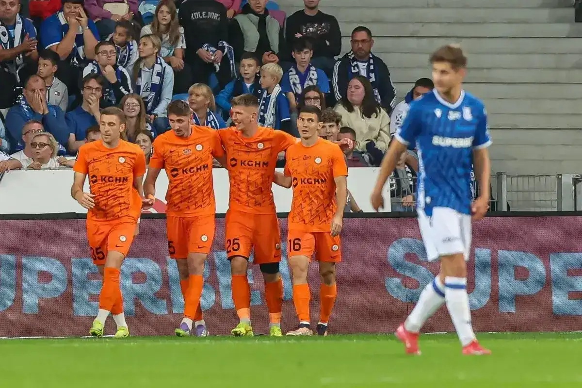 Piłkarze Zagłębia Lubin w pomarańczowych strojach świętują bramkę, podczas gdy zawodnik Lecha Poznań w niebieskim oddala się.