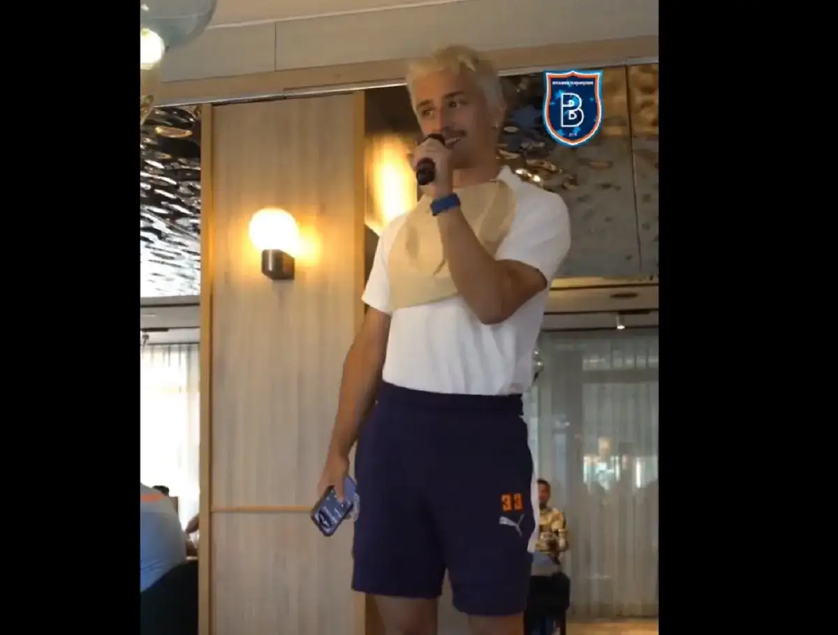 Młody mężczyzna z blond włosami, w koszulce i granatowych spodenkach z numerem 33, trzyma mikrofon. W tle logo İstanbul Başakşehir.