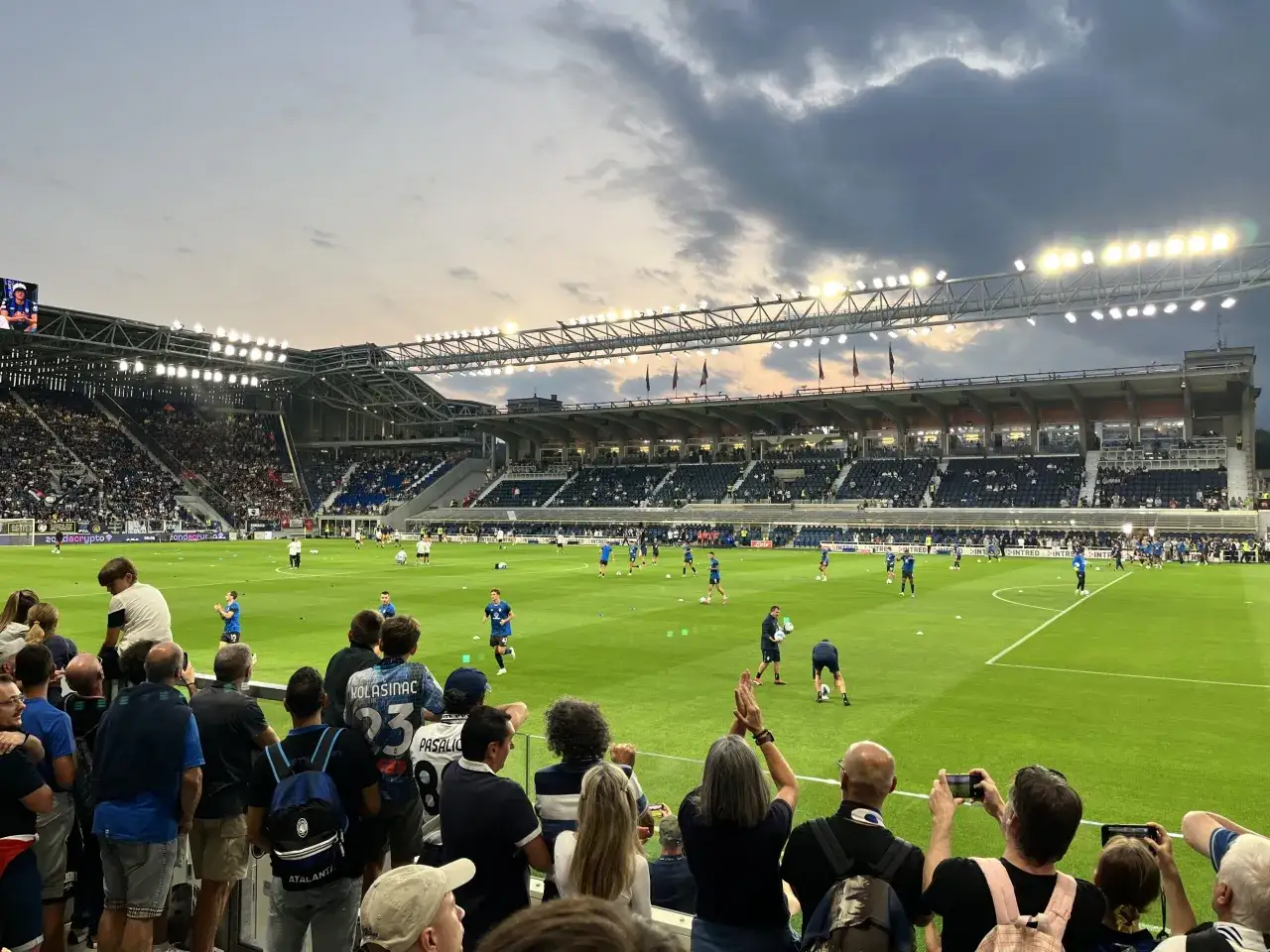 Piłkarze z Bergamo rozgrzewają się na murawie stadionu. Kibice na trybunach obserwują trening przed meczem.