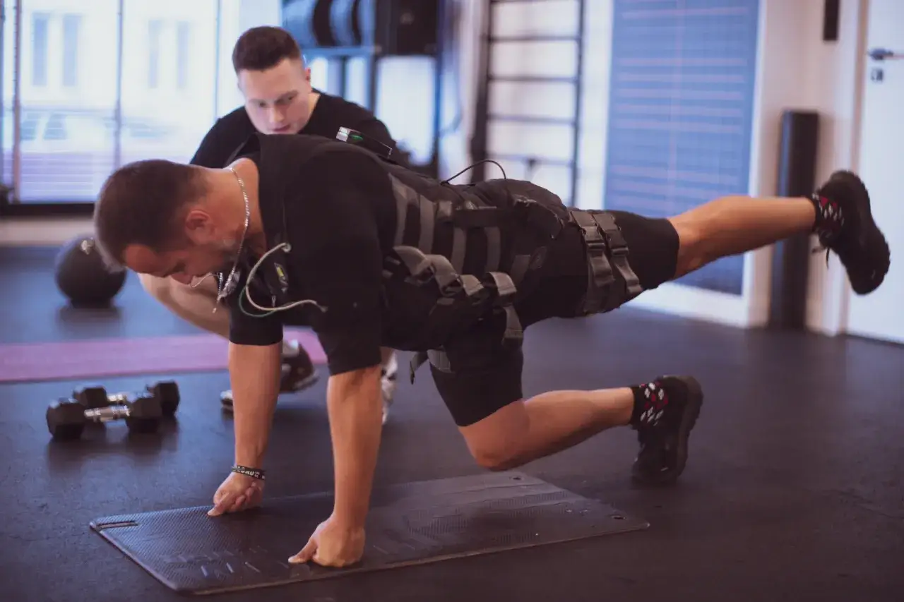 Trening EMS w 10 minut? Ten mężczyzna w kamizelce EMS wykonuje ćwiczenie, a trener go asekuruje. Czy 10 minutowe treningi dają efekty?