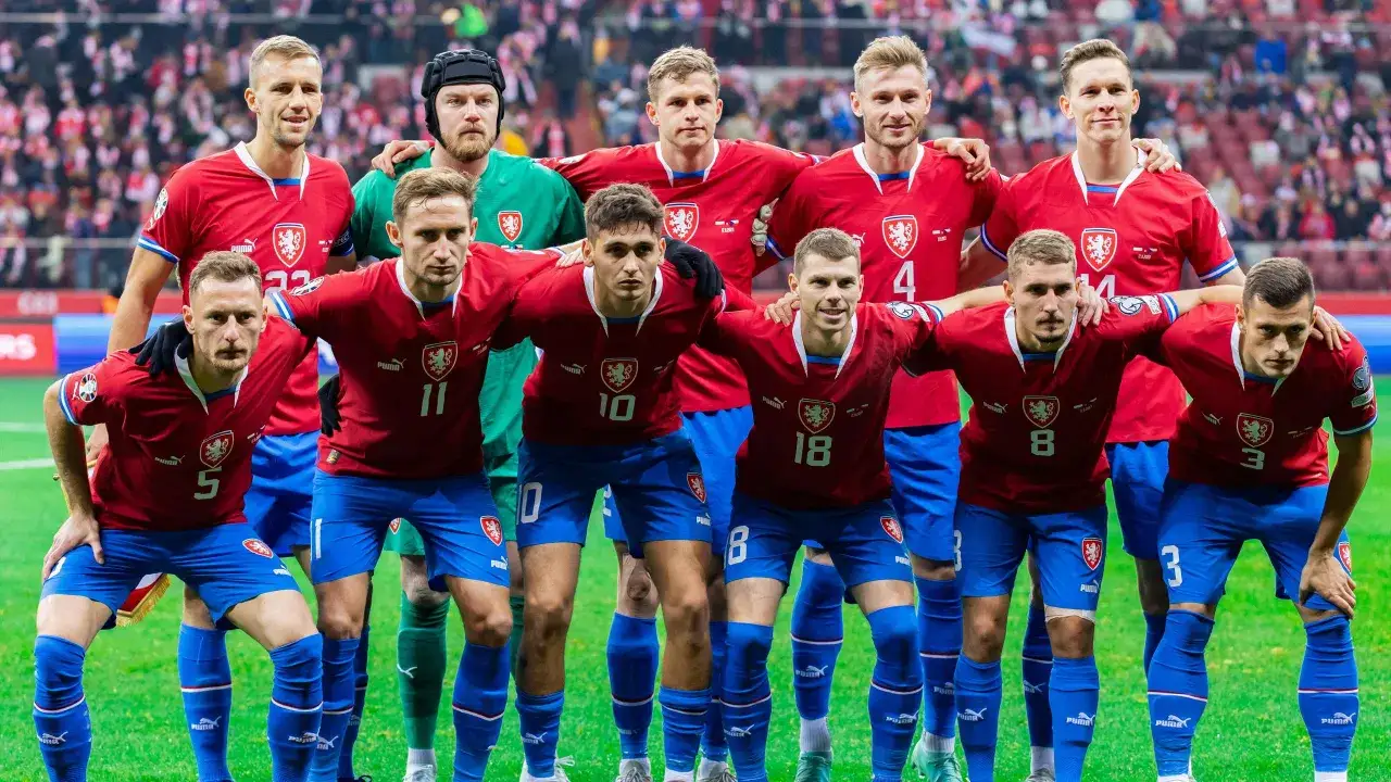 Drużyna piłkarska Czechów w czerwonych koszulkach i niebieskich spodenkach, gotowa do gry. Jeden z zawodników, czeski piłkarz, ma na głowie czarną czapkę bramkarską.