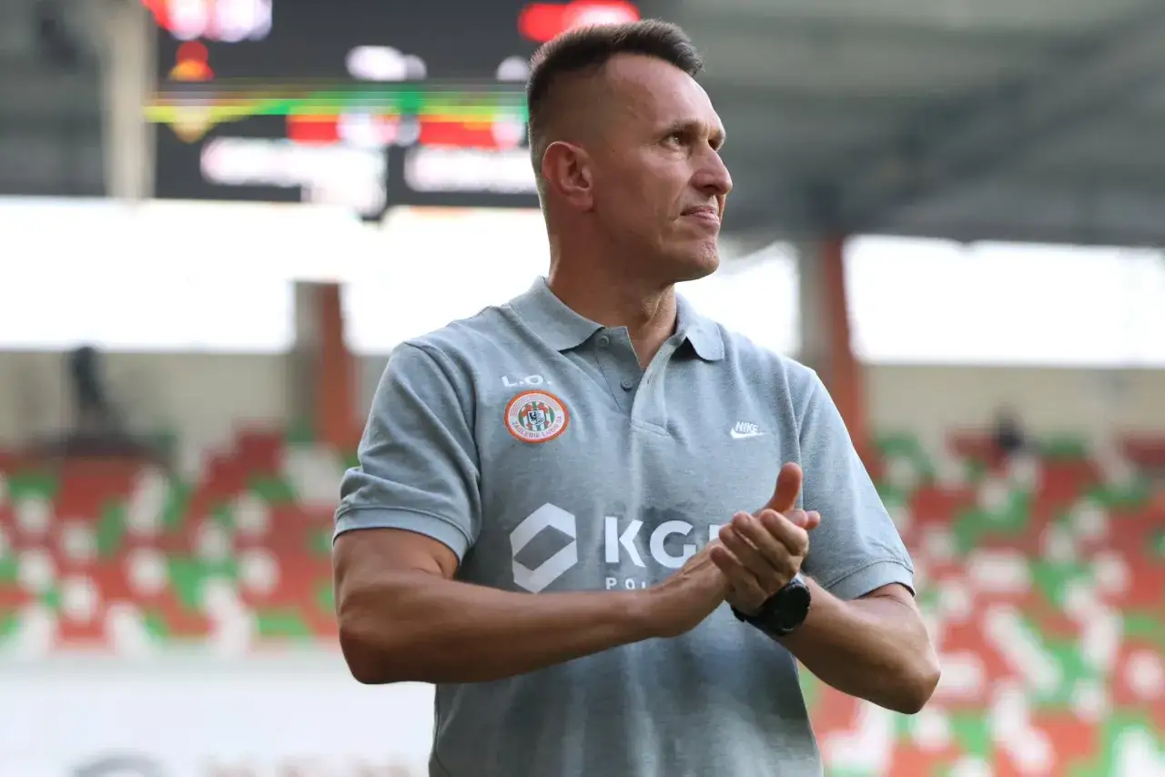 Trener Zagłębia Lubin, w szarej koszulce polo z herbem klubu, klaszcze na trybunach stadionu.