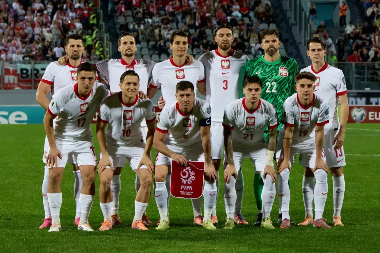 Drużyna Polski w piłce nożnej gotowa do gry. Piłkarze w białych strojach z orłem na piersi, na tle trybun.